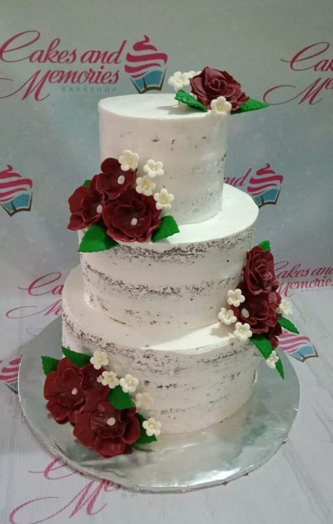 Three-tier semi-naked white cake decorated with deep red and white gumpaste flowers and green leaves.