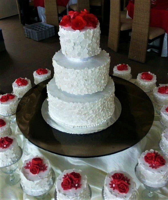 Elegant three-tier white wedding cake covered in white gumpaste flowers and topped with red roses, surrounded by matching mini cakes.