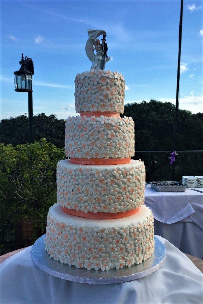 Four-tier white wedding cake covered in small white and peach fondant flowers with a bride and groom topper on a crescent moon.