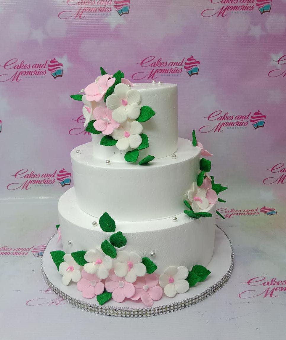 Elegant 3-tier white floral cake with pink and white gumpaste flowers and green leaves, accented with silver pearls.