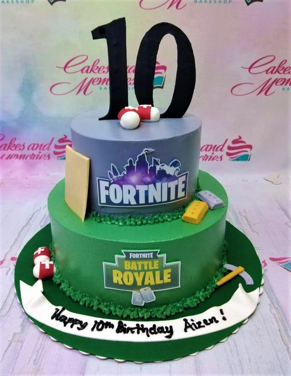 Two-tier Fortnite themed birthday cake with green and gray icing, featuring game logos, 3D fondant medkits, and a large black number 10 topper.