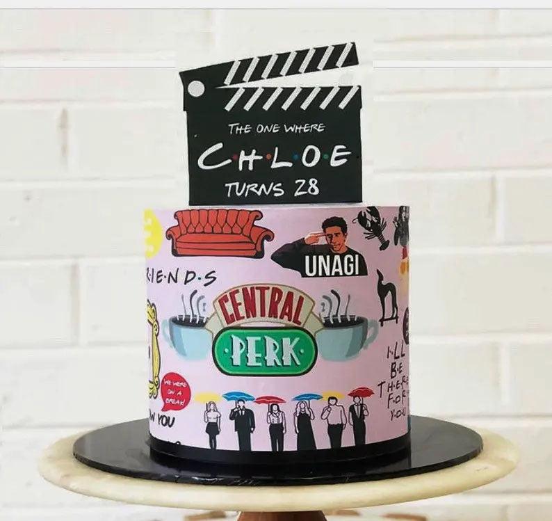 Friends TV show themed cake with a black clapperboard topper and a pink side wrap featuring iconic show symbols like the Central Perk logo and sofa.