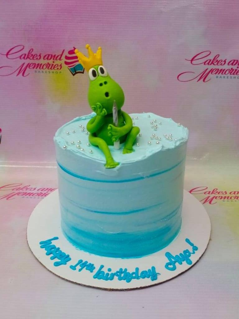 Light blue 1-tier birthday cake with a green frog figurine wearing a yellow crown and silver dragees.