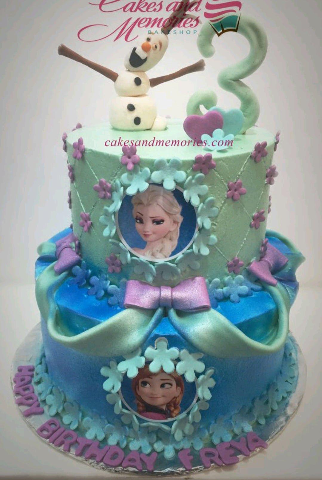 Two-tier Frozen themed cake in blue and mint green with Olaf topper, Elsa and Anna edible photos, and purple fondant bows.