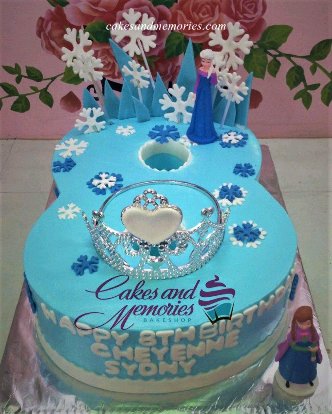 Blue number 8 shaped Frozen themed birthday cake with Elsa and Anna figurines, snowflakes, and a silver tiara.
