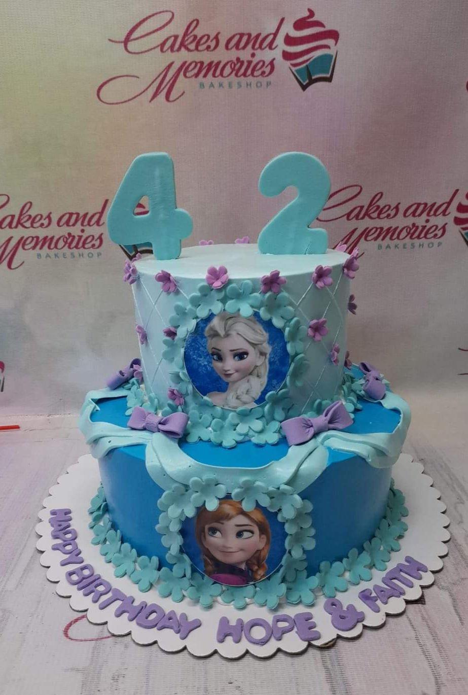 Two-tier Frozen themed birthday cake in blue and light blue with Elsa and Anna printouts, fondant flowers, and purple bows.