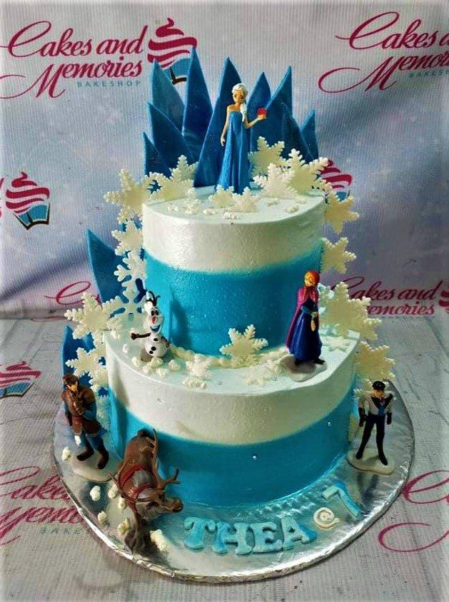 Two-tier Frozen themed birthday cake with Elsa, Anna, Olaf, Kristoff, and Sven figurines, white snowflakes, and blue mountain backdrops.