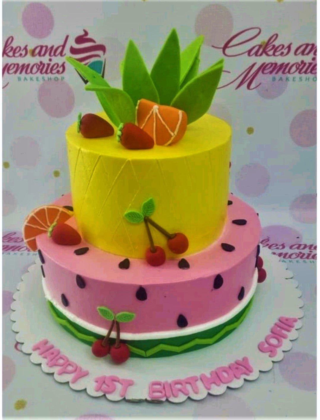 Two-tier fruit-themed birthday cake with yellow pineapple top tier and pink watermelon bottom tier featuring fondant fruit toppers.