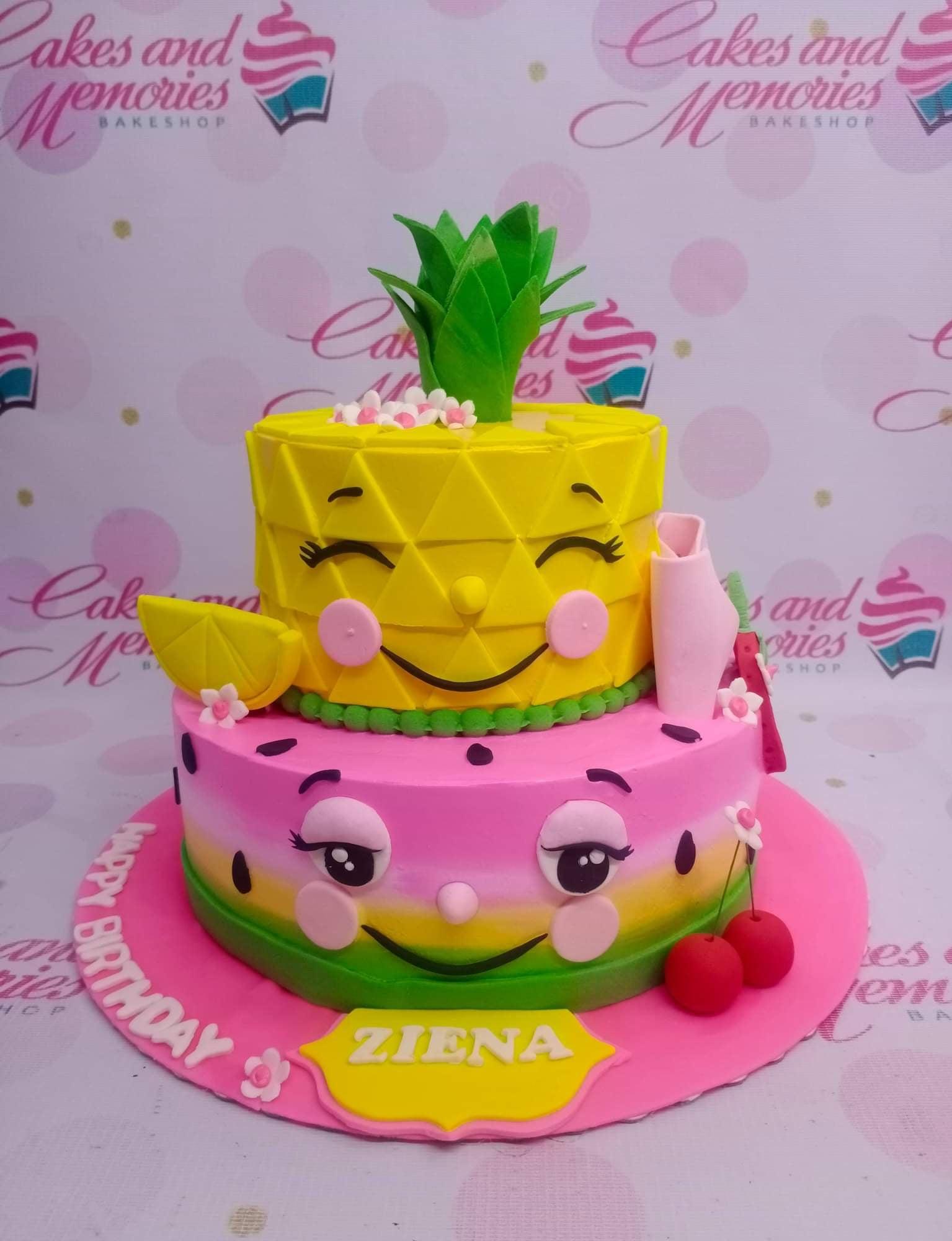 Two-tier fruit-themed fondant cake featuring a yellow pineapple top tier and a pink watermelon bottom tier with cute faces and fruit accents.