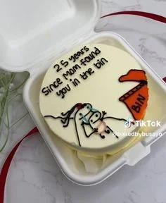 White bento cake with a funny stick figure drawing of a mom finding a baby in a bin and a humorous birthday message.
