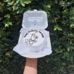 White bento cake with a hand-drawn doodle of a character sitting on a toilet wearing a crown and holding a small cake.