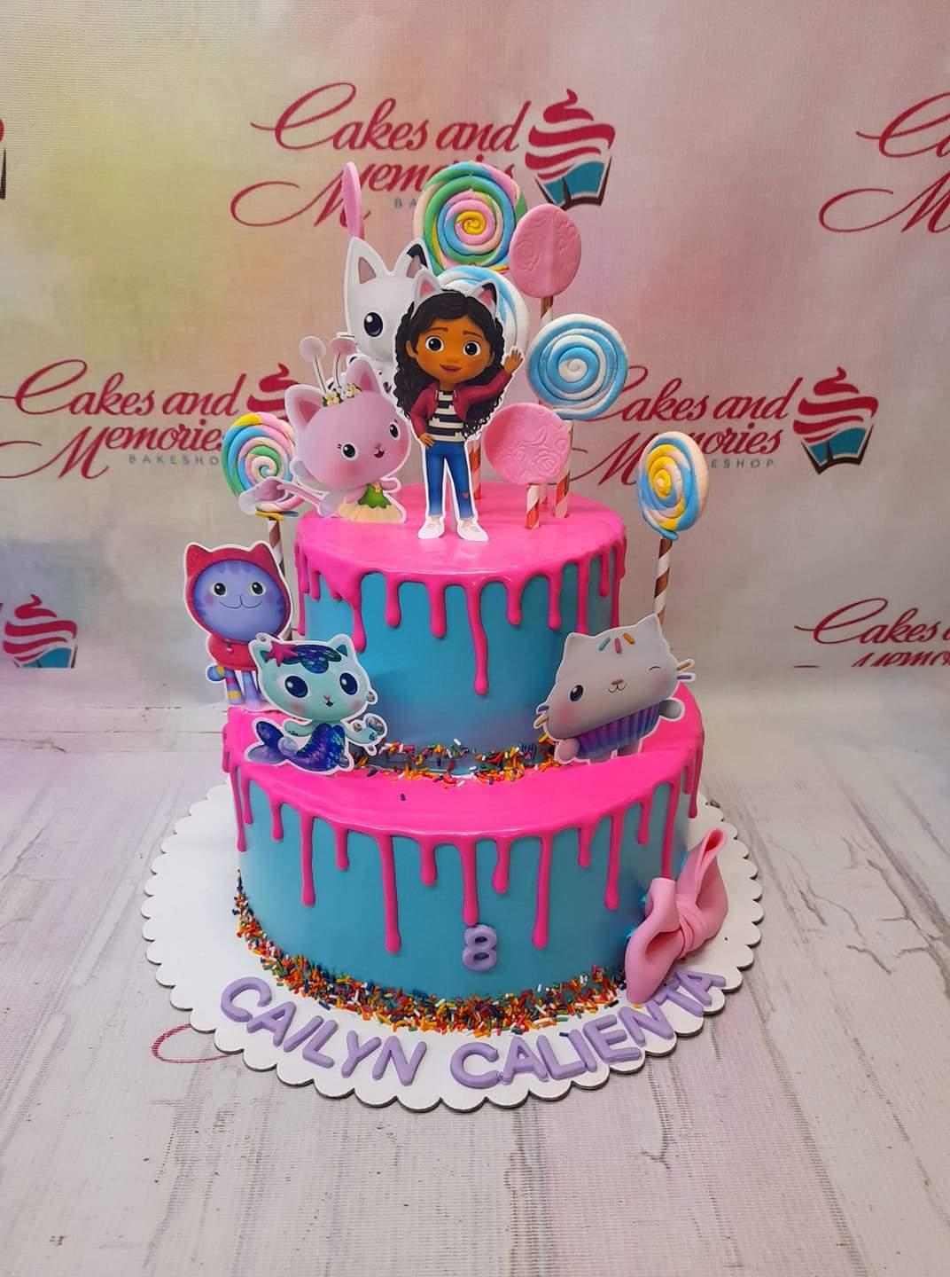 Two-tier Gabby's Dollhouse themed cake in light blue and hot pink with character printouts, lollipops, and colorful sprinkles.