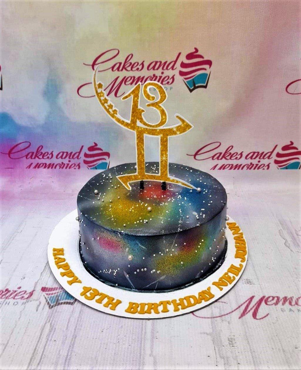 A single-tier galaxy-themed birthday cake with a dark blue and black airbrushed finish, silver dragees, and a gold glitter number 13 topper.
