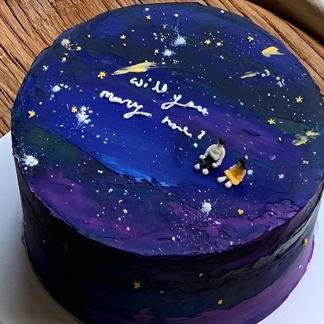 Galaxy themed bento cake with purple and navy blue icing, white stars, and a miniature couple figurine with a proposal message.