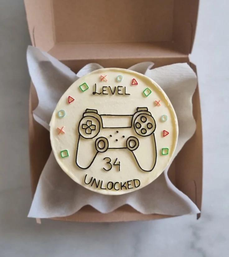 Ivory bento cake with a hand-drawn black icing game controller and colorful PlayStation symbols for a 34th birthday.