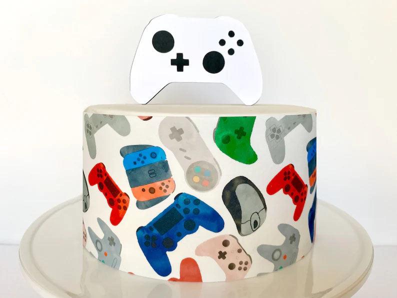 White single tier cake with a gaming theme featuring a side wrap of colorful video game controllers and a white controller topper.
