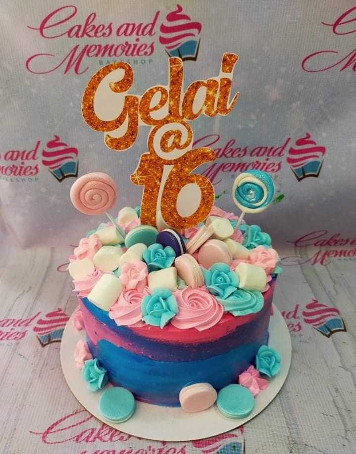 Pink and blue 16th birthday cake with glittery gold name topper, icing rosettes, marshmallows, and macarons.