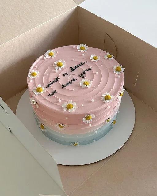 Pink and blue gradient minimalist cake with white daisy flowers and a gender reveal message in black icing.