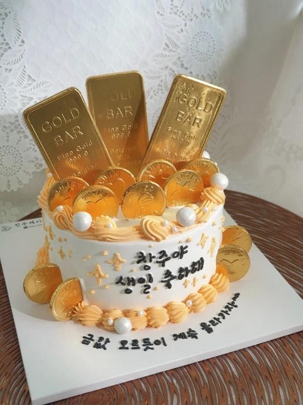 White soft-icing cake decorated with gold chocolate bars, gold coins, and white pearls with Korean birthday message.