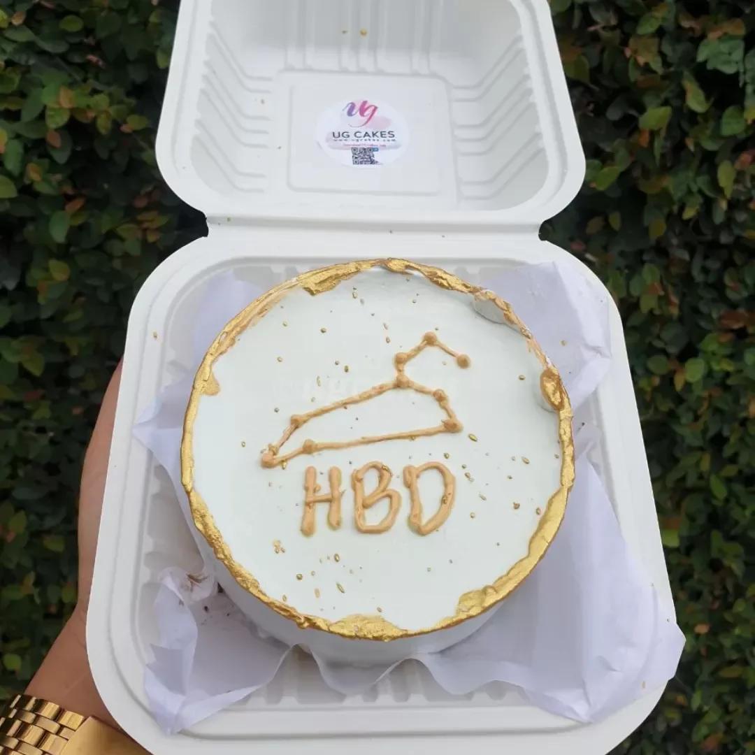White bento cake with gold painted edges and a gold constellation design with HBD message in a white takeout box.