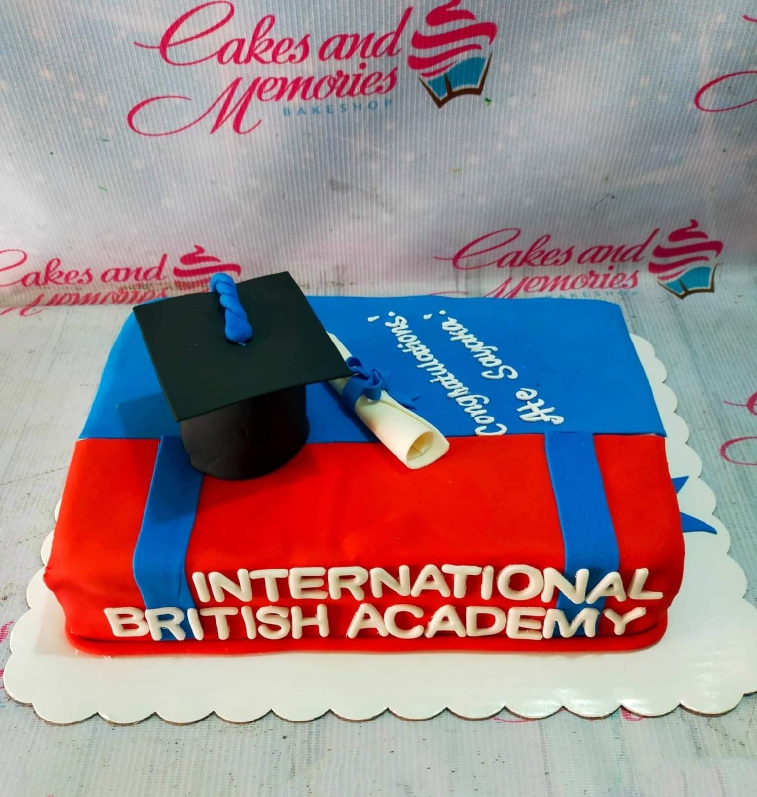 Rectangle graduation cake with red and blue fondant, featuring a black graduation cap, a white diploma, and the text International British Academy.