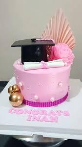 Pink graduation cake with a black graduation cap, white diploma, pink paper fan, and gold spheres on a white base board.