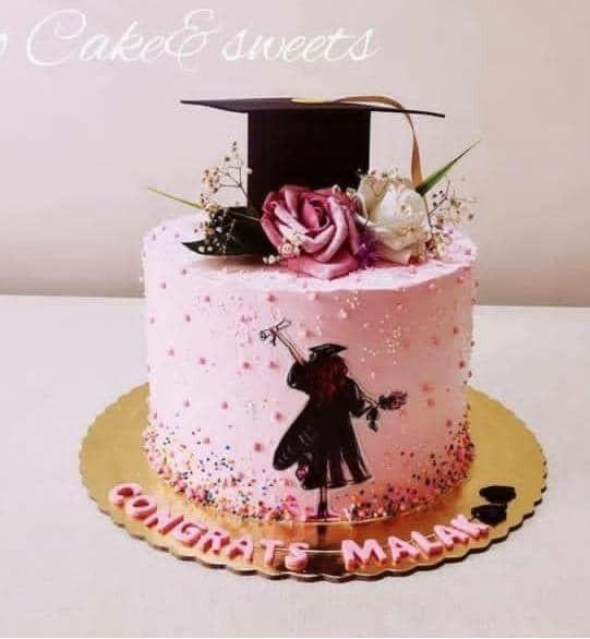 Pink graduation cake with a black mortarboard topper, pink and white roses, and a silhouette of a graduate holding a diploma.