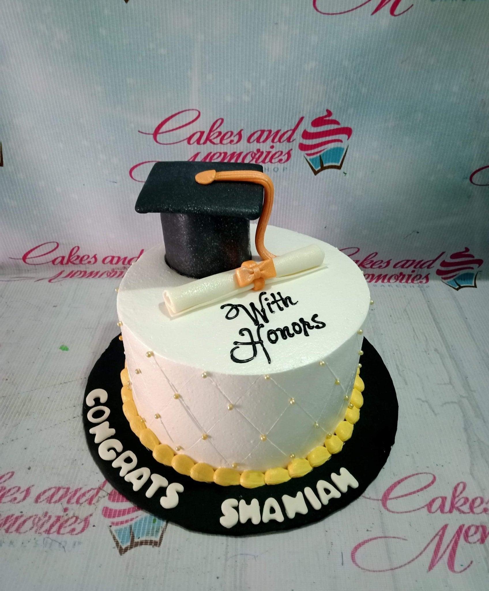 White graduation cake with a black fondant graduation cap, a white diploma with an orange bow, and gold pearl sprinkles.