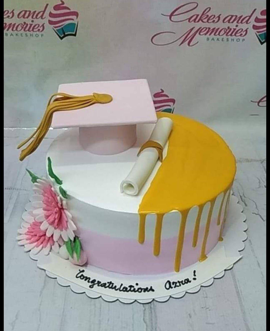 White and pink graduation cake with a pink graduation cap, white diploma, and pink flowers with yellow drip icing.