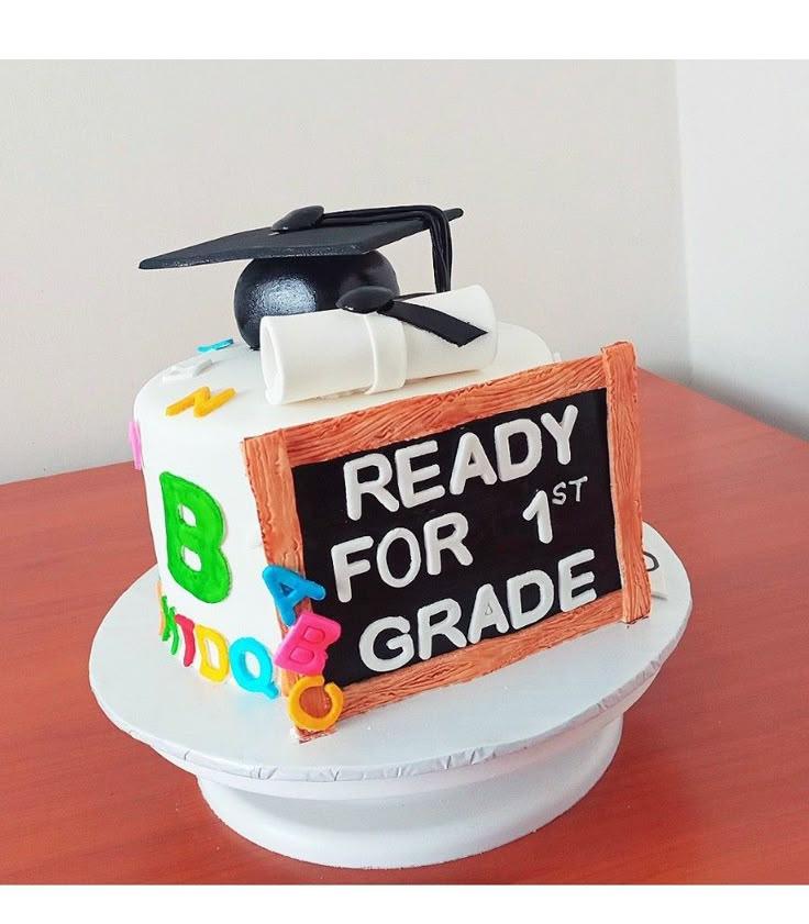 White fondant graduation cake with a black graduation cap, diploma, and a chalkboard saying Ready for 1st Grade with colorful alphabet letters.