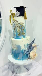Two-tier white and blue watercolor graduation cake with a black graduation cap, diploma, and gold Class of 2024 topper.