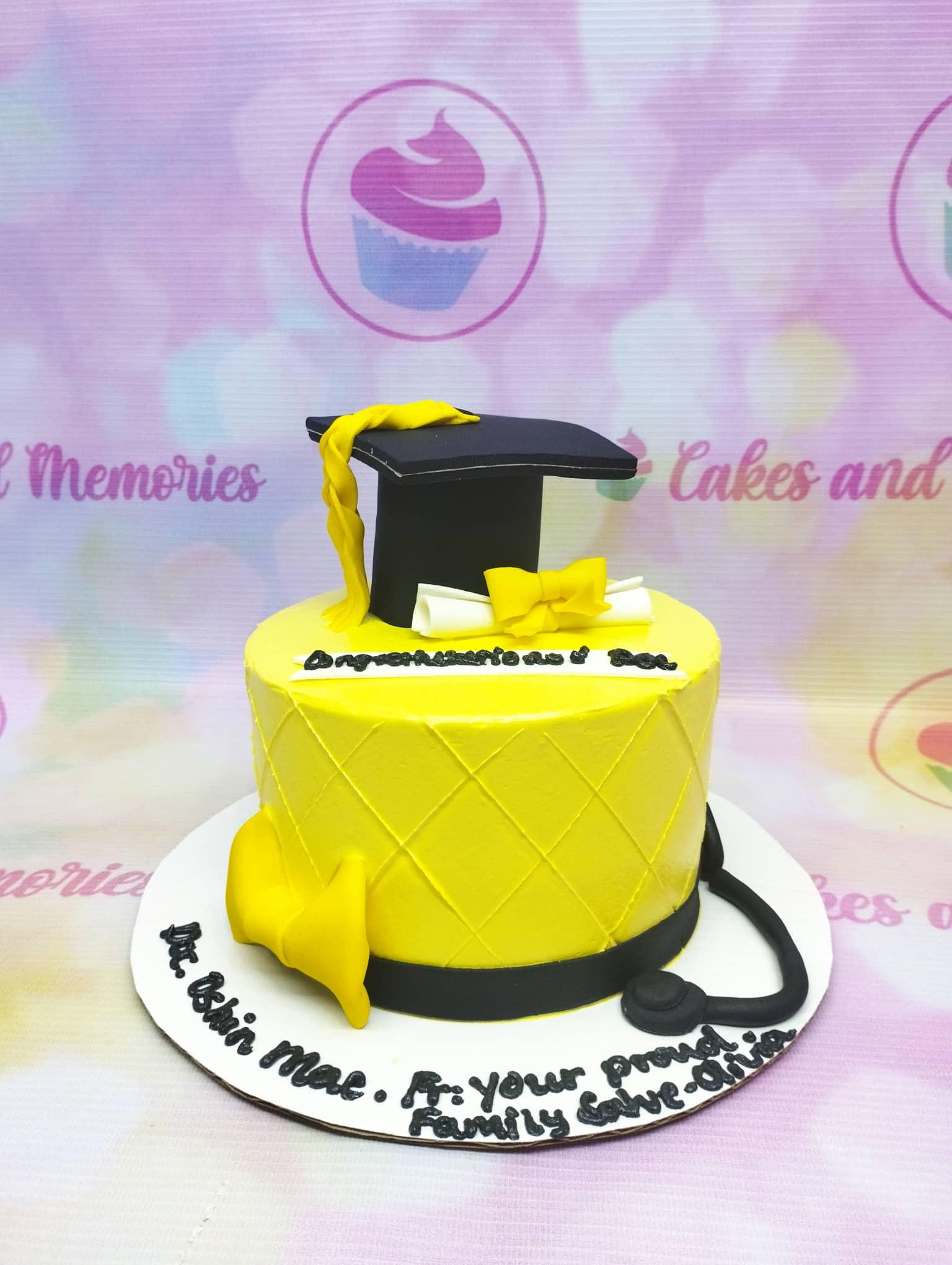 Yellow graduation cake with a black fondant graduation cap, diploma, and stethoscope on a white base board.