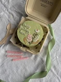 A light green bento cake with white piped birthday message and delicate pink and white icing flowers in a beige box.
