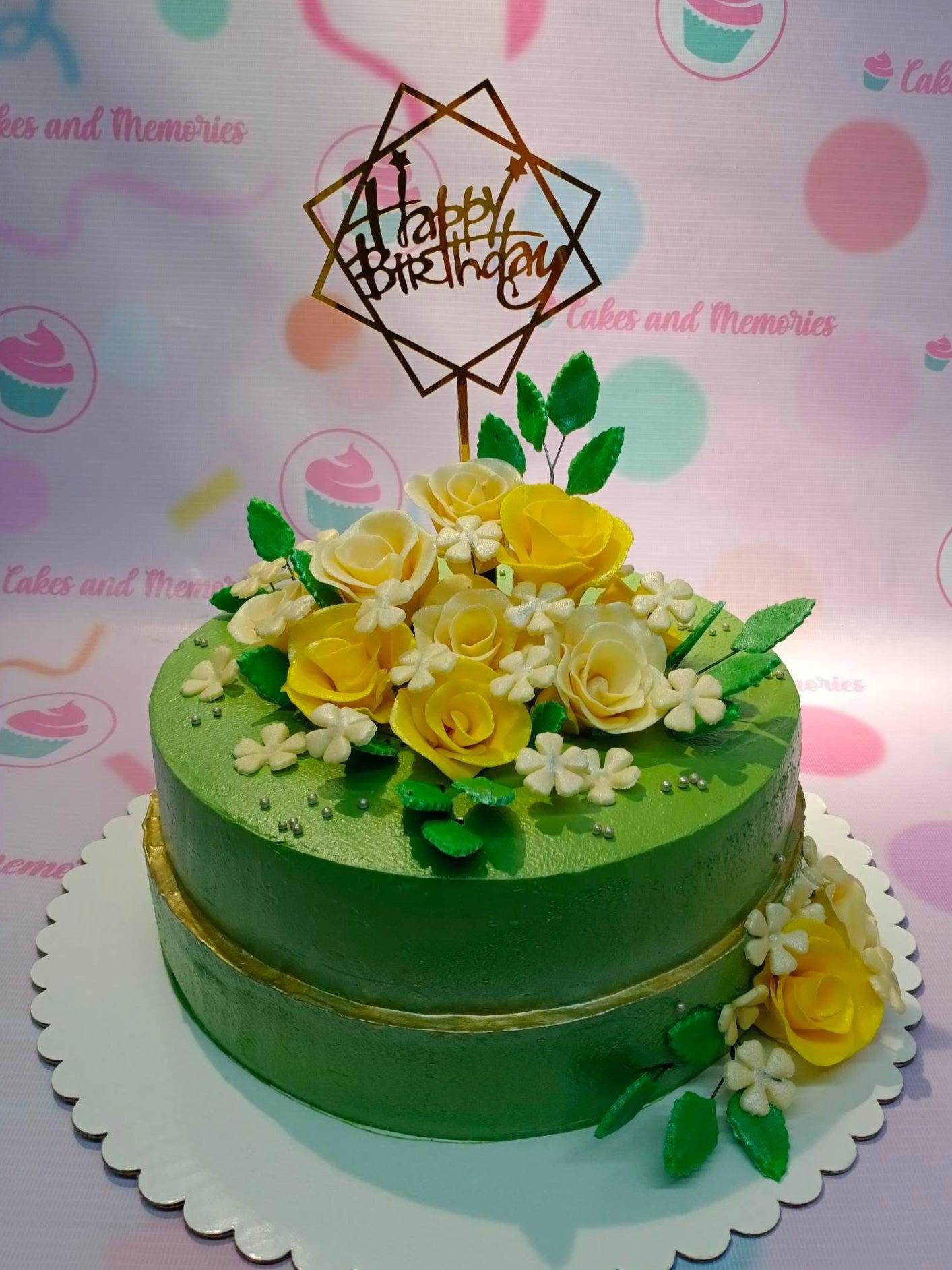 Green one-tier birthday cake decorated with yellow and white gumpaste roses, green leaves, and a gold geometric Happy Birthday topper.