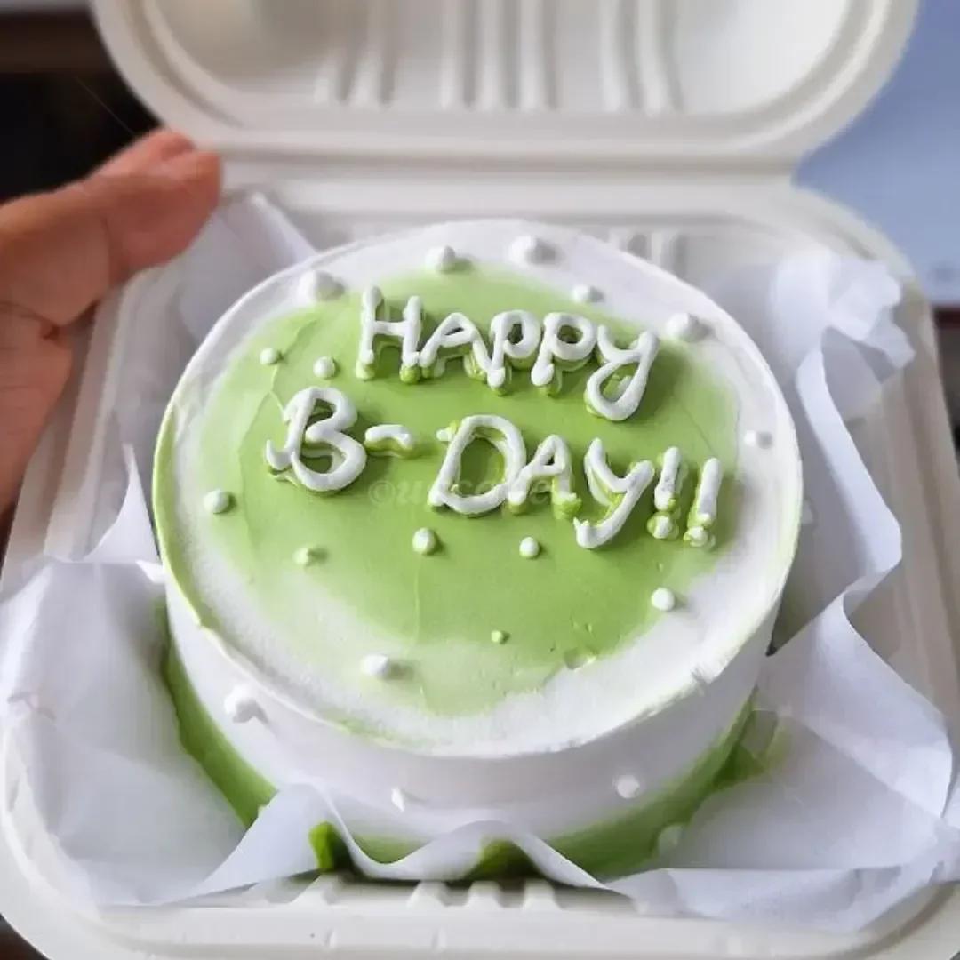 Green and white minimalist bento cake with piped icing message Happy B-Day and white icing dots in a white container.