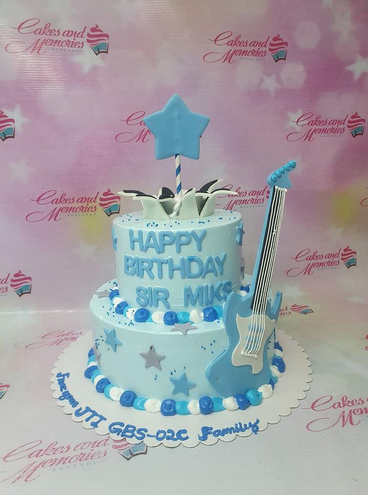 Two-tier light blue birthday cake with a blue guitar topper, fondant stars, and blue and white icing dollops.