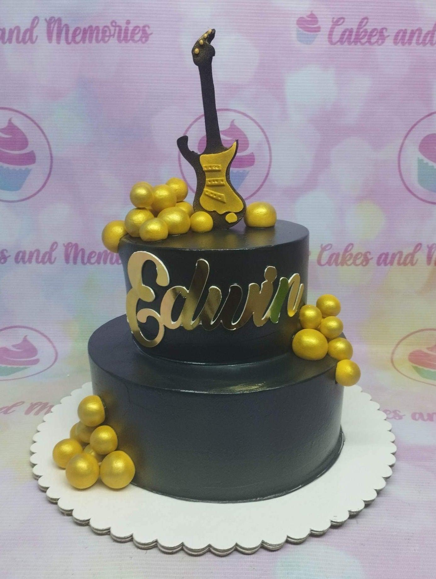 Two-tier black cake with a gold guitar topper, gold spheres, and a gold name cutout.