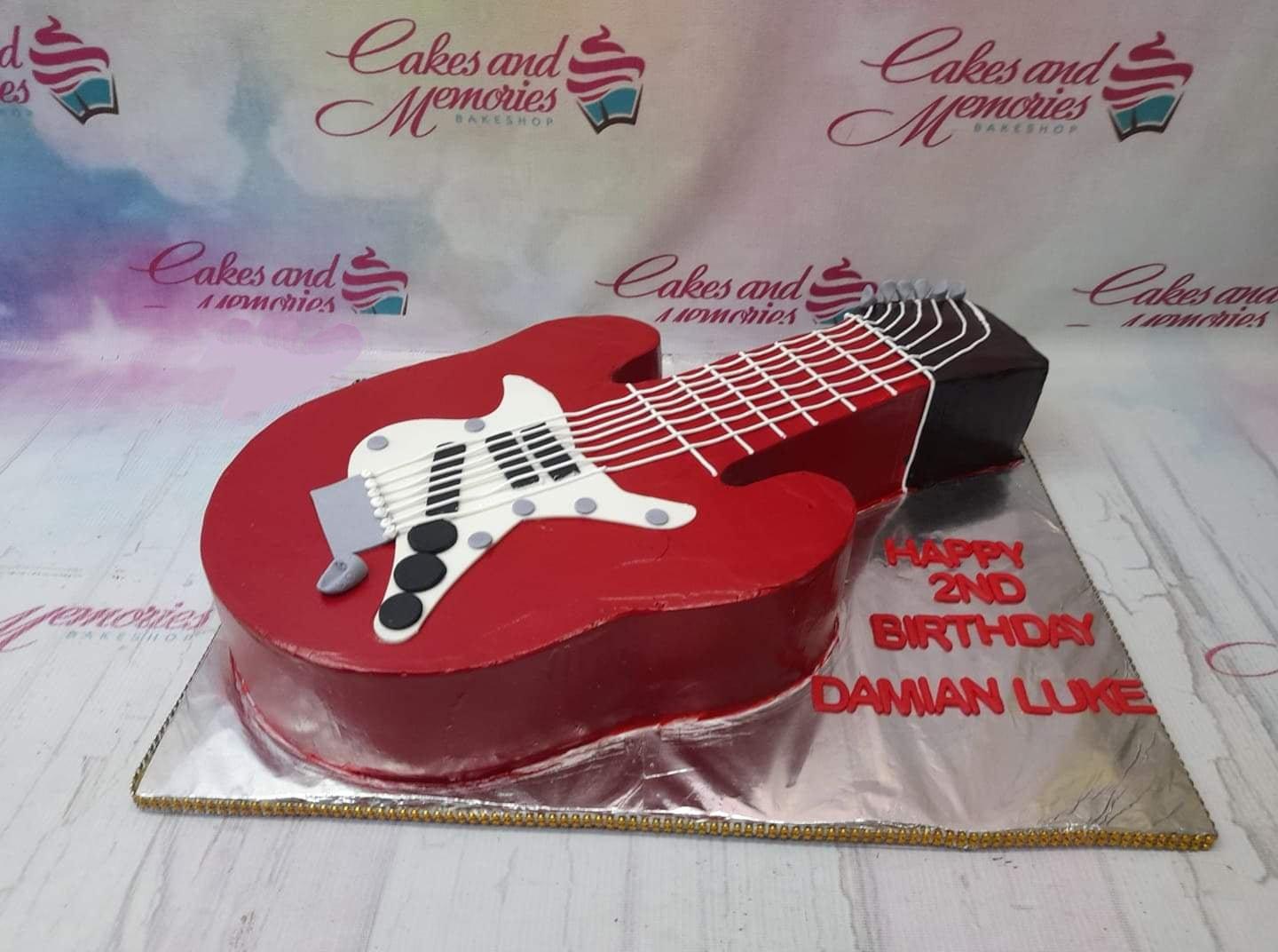 Red electric guitar shaped cake with white pickguard and black knobs on a silver board for a 2nd birthday celebration.