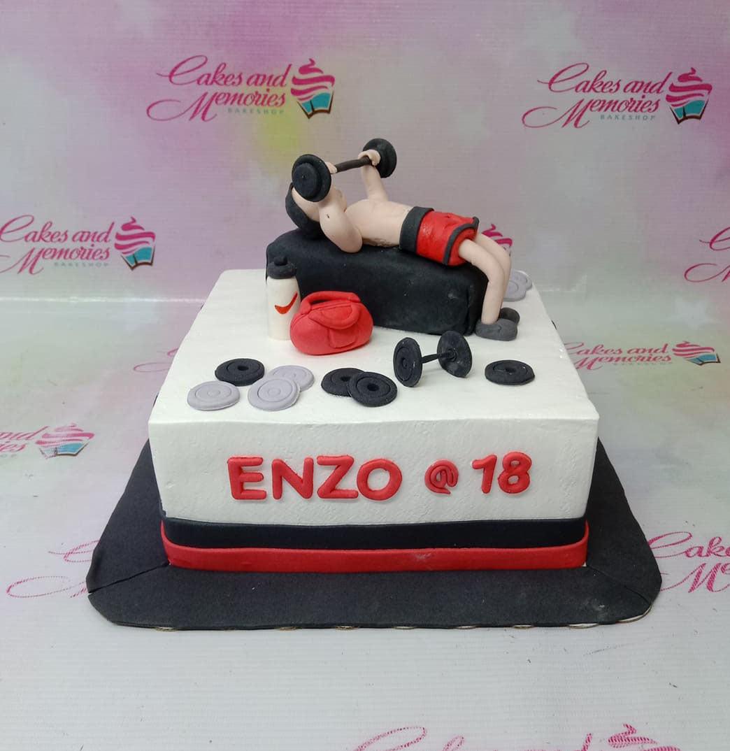 Square white gym themed cake with a fondant man lifting weights on a bench, red gym bag, and dumbbells.