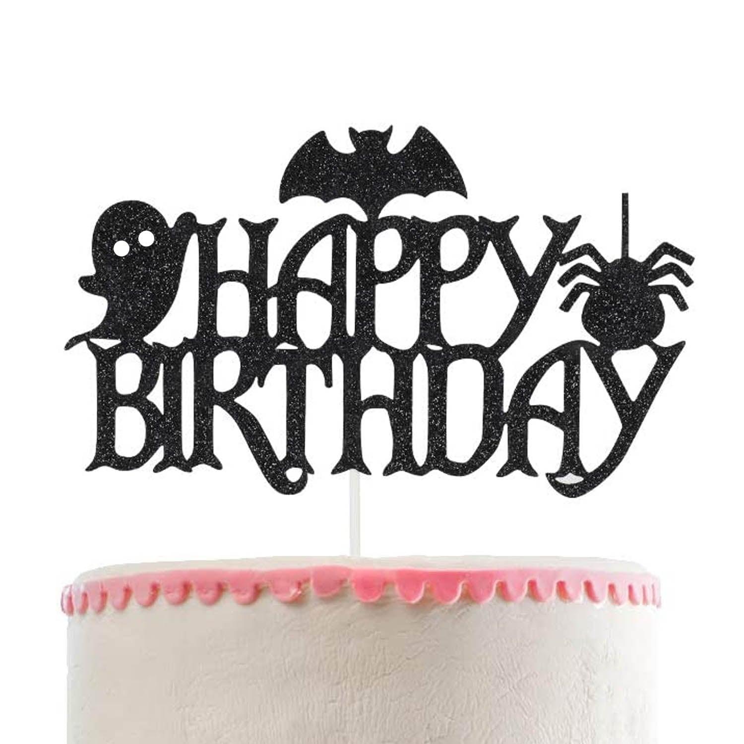 White soft-icing cake with a pink piped border and a black glittery Halloween-themed Happy Birthday topper featuring a bat, ghost, and spider.