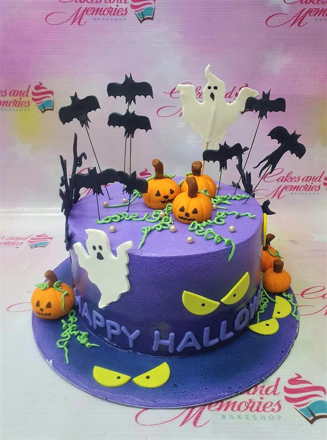 Purple Halloween cake with fondant pumpkins, ghosts, black bat toppers, and yellow spooky eyes on a purple base board.