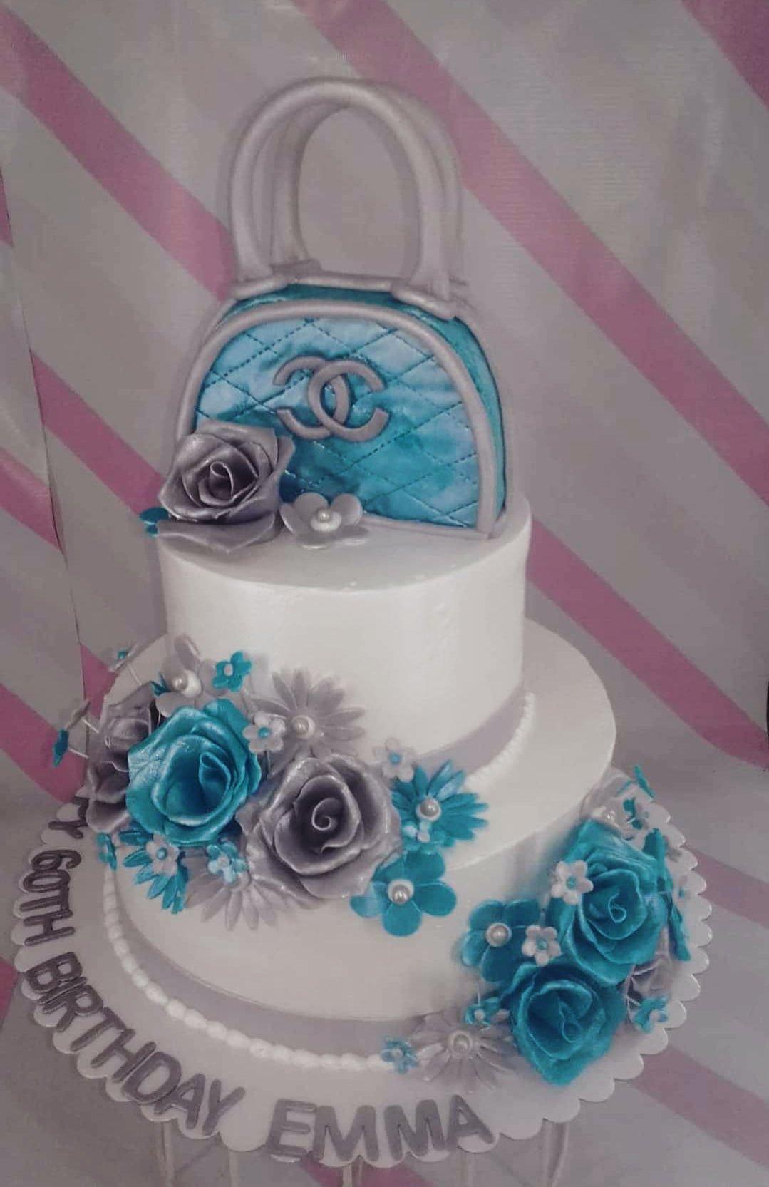 Two-tier white fondant cake with blue and silver roses, featuring a blue quilted handbag topper with a silver logo.