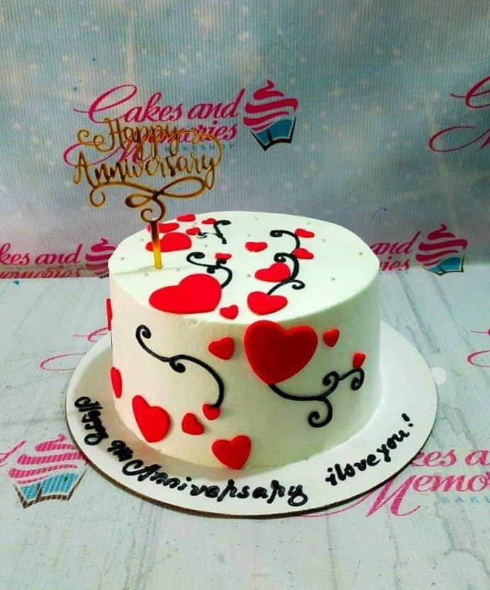 White anniversary cake with red fondant hearts and black icing swirls, topped with a gold Happy Anniversary topper.
