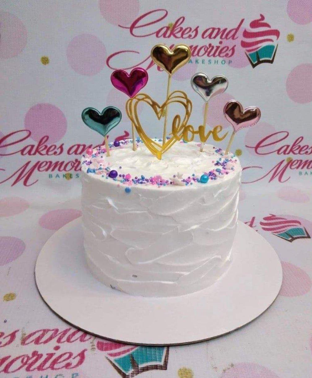 White soft-icing cake with textured palette knife finish, colorful heart toppers, and purple and pink sprinkles.