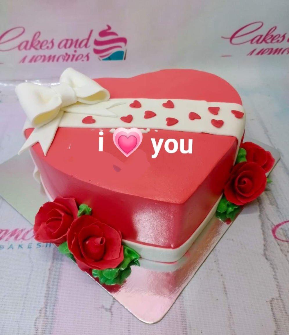 Heart-shaped red cake with white fondant bow and ribbon, red roses, and 'i love you' message.