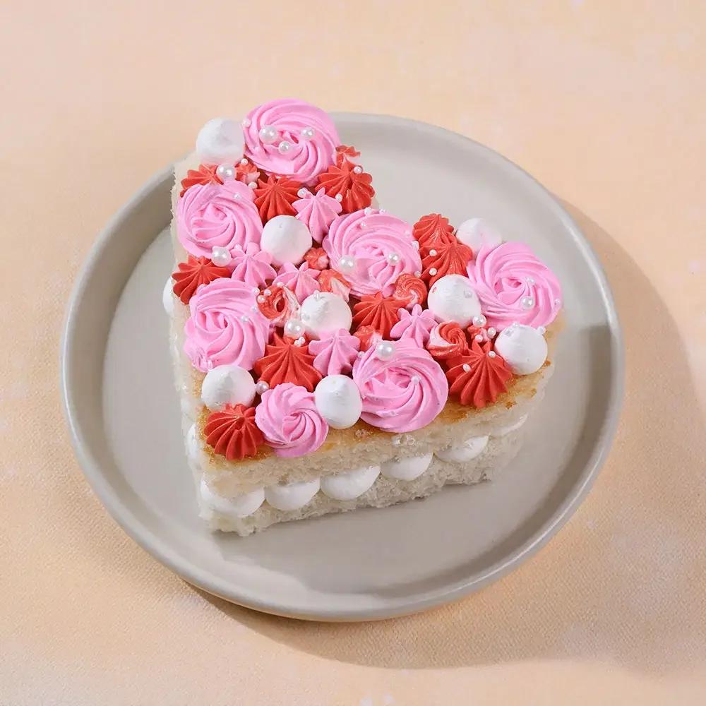 Heart-shaped bento cake with pink and red icing rosettes, white meringue kisses, and pearl sprinkles on a cream plate.