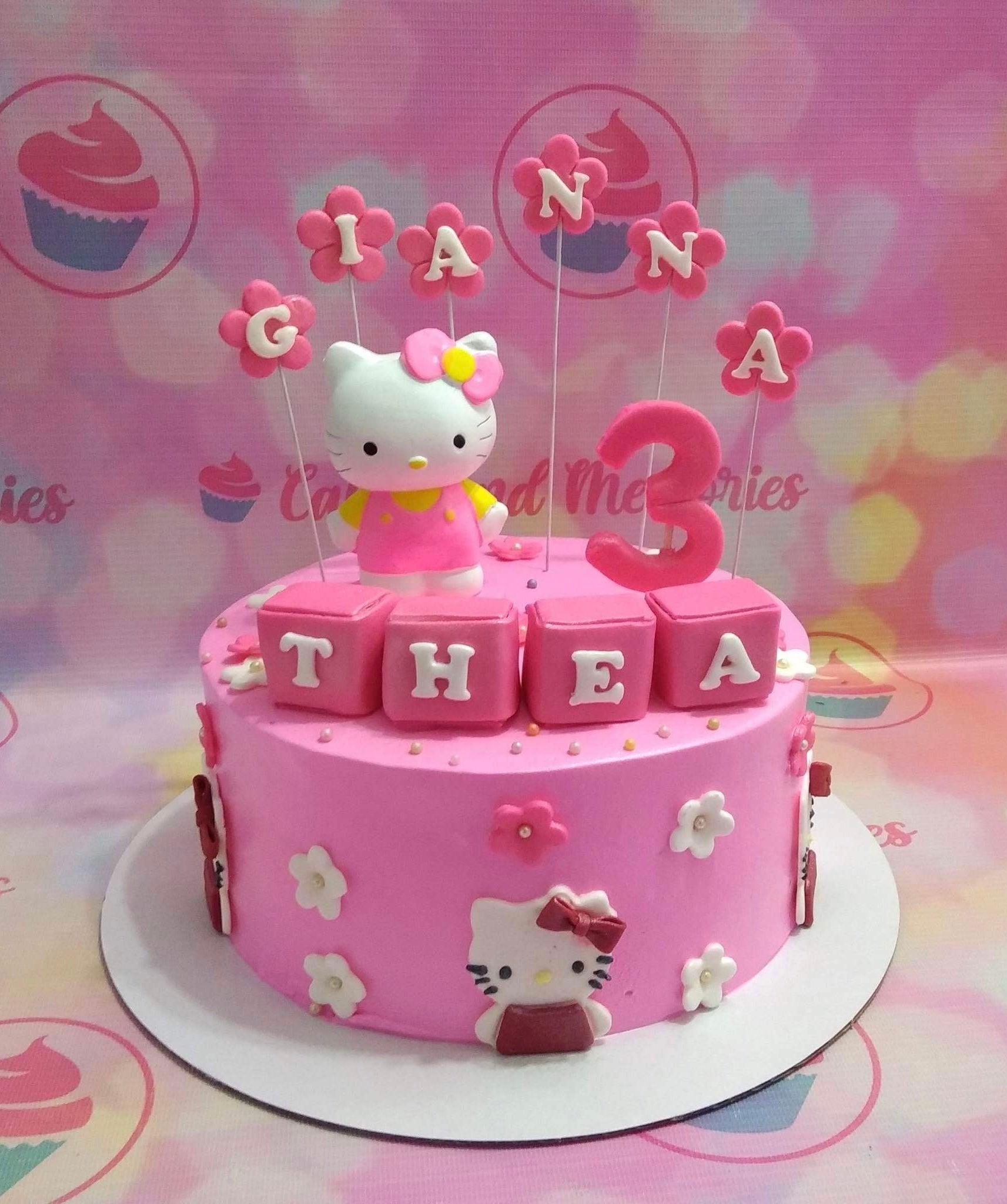 Pink Hello Kitty themed birthday cake with a 3D Hello Kitty figure, pink flower toppers, and name blocks for Thea's 3rd birthday.