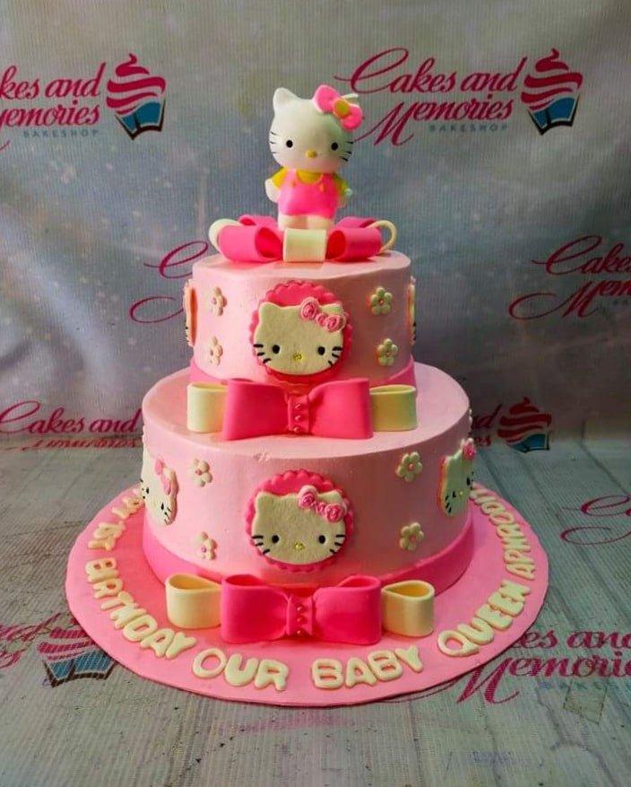 Two-tier pink Hello Kitty fondant cake with pink bows, edible flowers, and a 3D Hello Kitty topper on a pink base board.