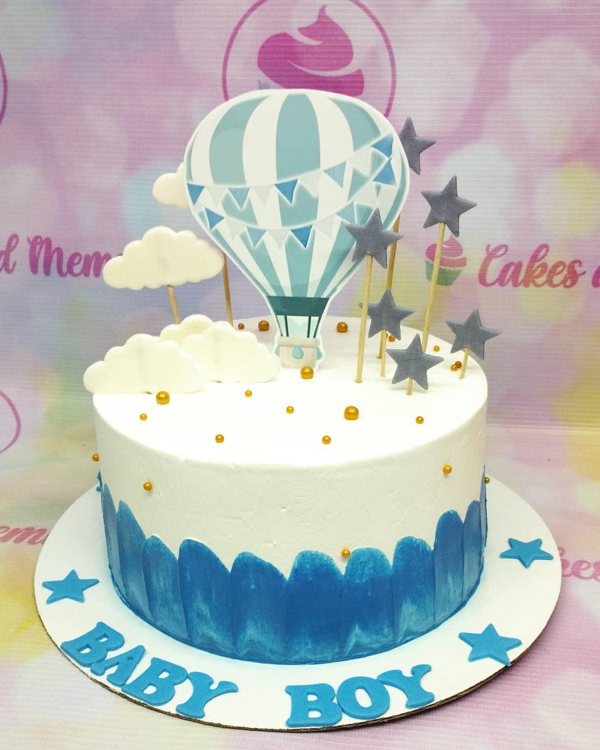 White 1-tier baby boy cake with blue hot air balloon printout, white fondant clouds, and gray stars.