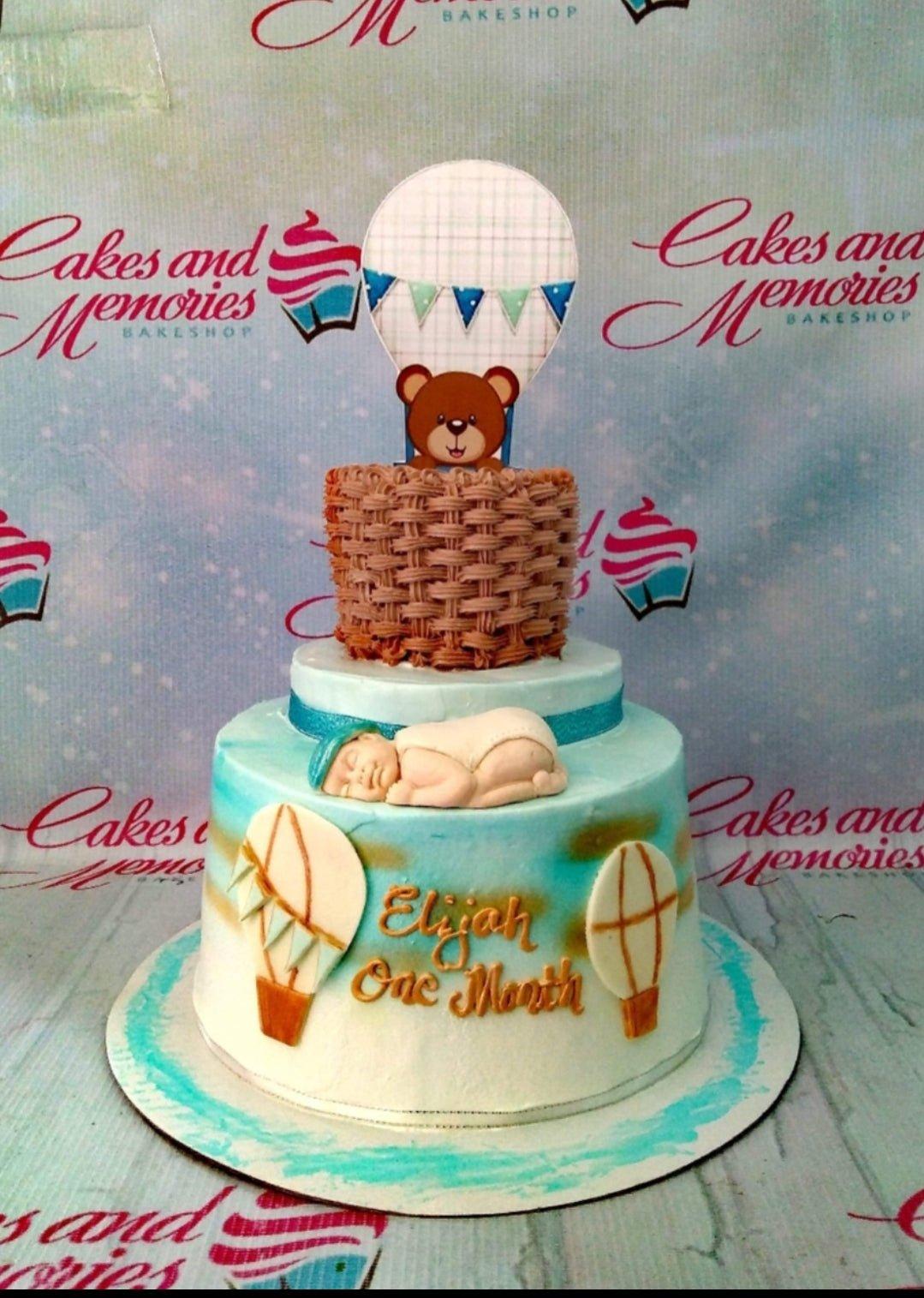 Two-tier light blue baby boy cake with a sleeping baby fondant figure, hot air balloon toppers, and a brown bear in a basket.